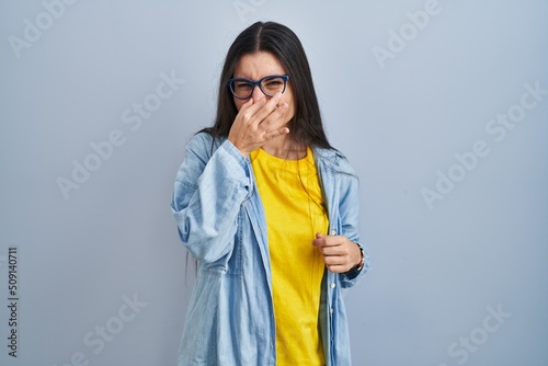 Young hispanic woman standing over blue background smelling something stinky and disgusting, intolerable smell, holding breath with fingers on nose. bad smell