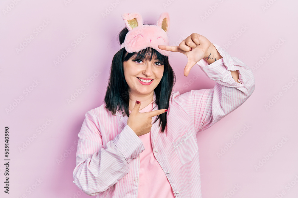 Young hispanic woman wearing sleep mask and pajama smiling making frame with hands and fingers with happy face. creativity and photography concept.