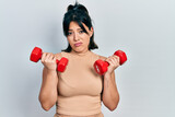 Young hispanic woman wearing sportswear using dumbbells clueless and confused expression. doubt concept.
