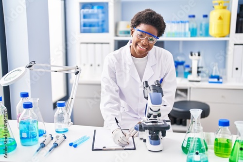 African american woman wearing scientist uniform using microscope write on clipboard at laboratory