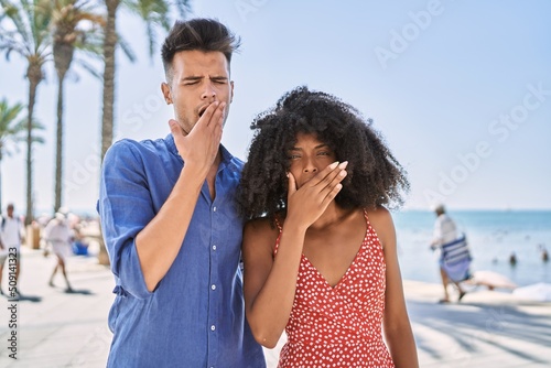 Young interracial couple outdoors on a sunny day bored yawning tired covering mouth with hand. restless and sleepiness.