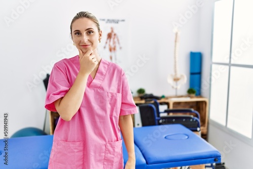 Young blonde woman working at pain recovery clinic looking confident at the camera with smile with crossed arms and hand raised on chin. thinking positive.