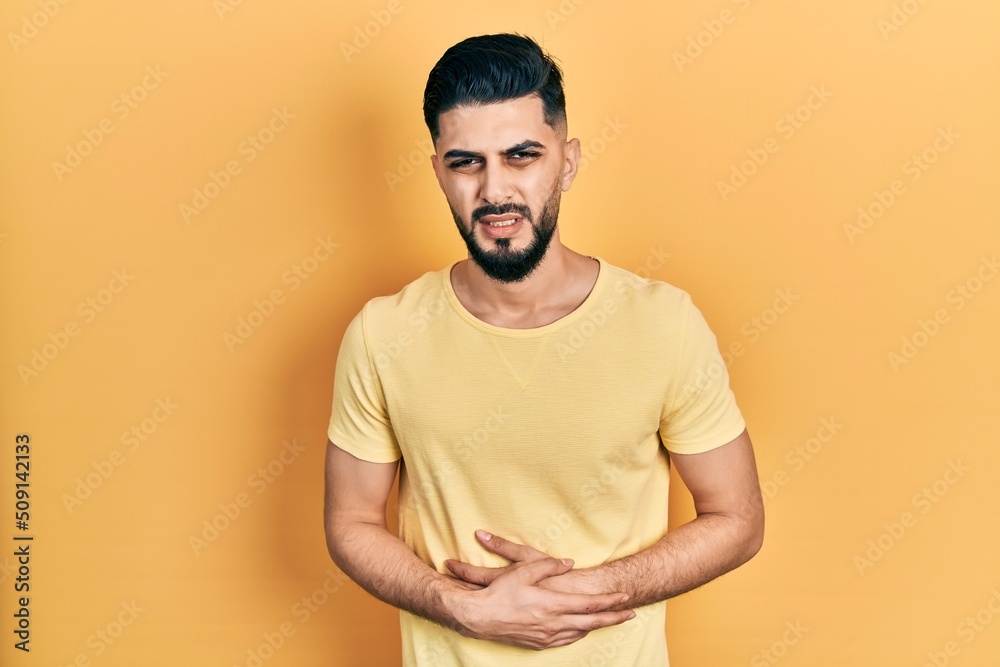 Handsome man with beard wearing casual yellow t shirt with hand on stomach because indigestion, painful illness feeling unwell. ache concept. Handsome man with beard wearing casual yellow t shirt with hand on stomach because indigestion, painful illness feeling unwell. ache concept.