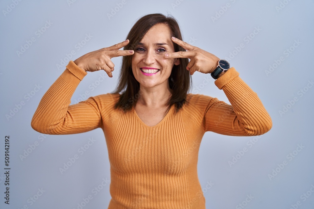 Middle age brunette woman standing wearing orange sweater doing peace symbol with fingers over face, smiling cheerful showing victory Middle age brunette woman standing wearing orange sweater doing peace symbol with fingers over face, smiling cheerful showing victory