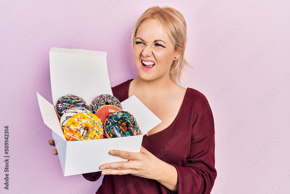 Young blonde woman holding tasty colorful doughnuts angry and mad screaming frustrated and furious, shouting with anger. rage and aggressive concept. Young blonde woman holding tasty colorful doughnuts angry and mad screaming frustrated and furious, shouting with anger. rage and aggressive concept.