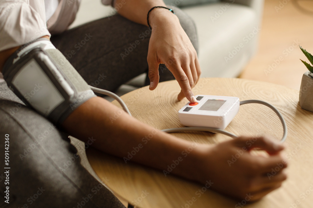 Fototapeta premium Cropped view of millennial black man sitting on couch at home, checking blood pressure, using modern tonometer