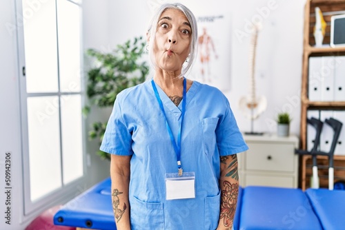 Middle age grey-haired woman wearing physiotherapist uniform at medical clinic making fish face with lips, crazy and comical gesture. funny expression.