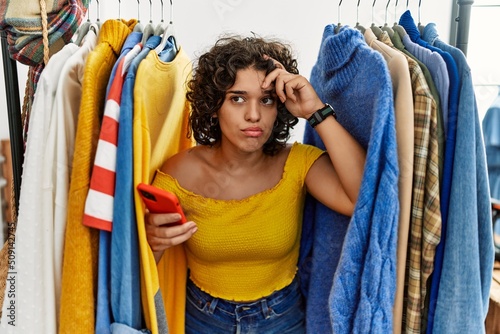 Young hispanic woman searching clothes on clothing rack using smartphone worried and stressed about a problem with hand on forehead, nervous and anxious for crisis