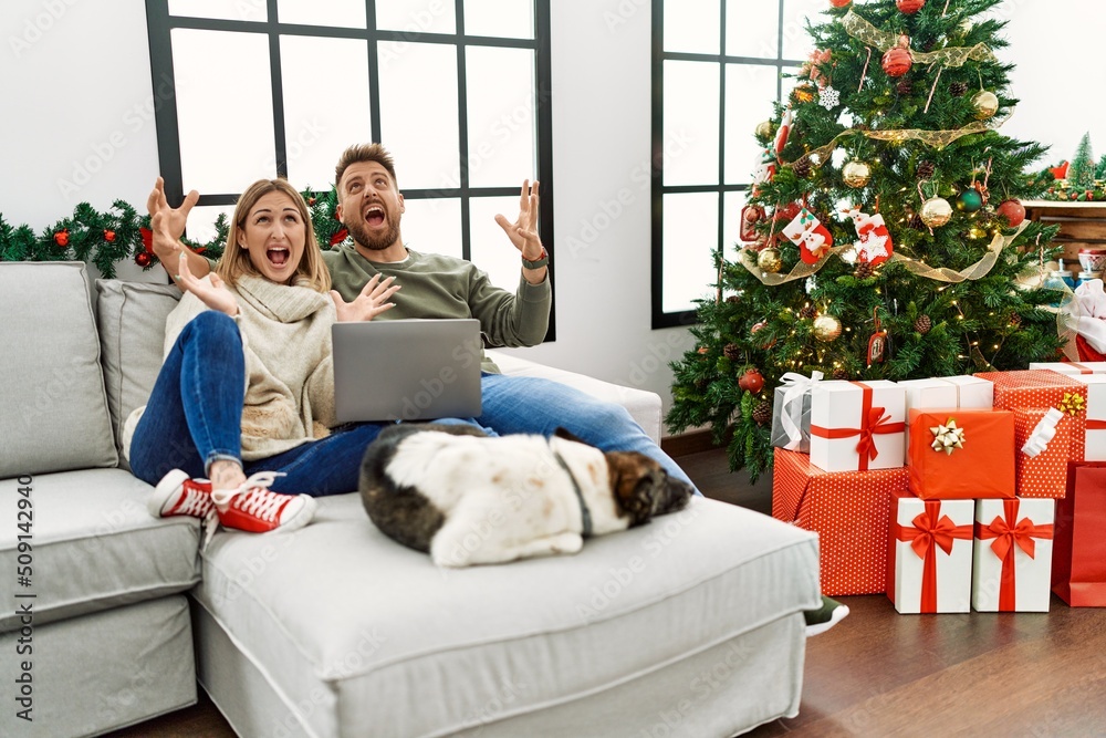 Young couple using laptop sitting by christmas tree crazy and mad shouting and yelling with aggressive expression and arms raised. frustration concept. Young couple using laptop sitting by christmas tree crazy and mad shouting and yelling with aggressive expression and arms raised. frustration concept.