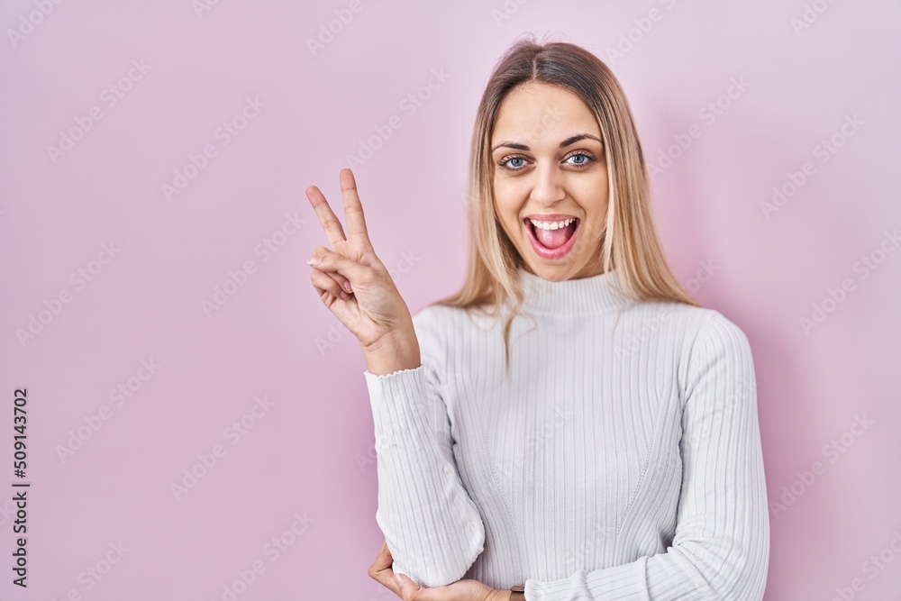 Young blonde woman wearing white sweater over pink background smiling with happy face winking at the camera doing victory sign. number two. Young blonde woman wearing white sweater over pink background smiling with happy face winking at the camera doing victory sign. number two.