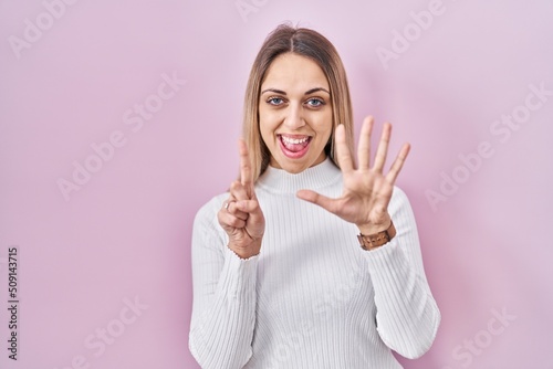 Young blonde woman wearing white sweater over pink background showing and pointing up with fingers number six while smiling confident and happy.