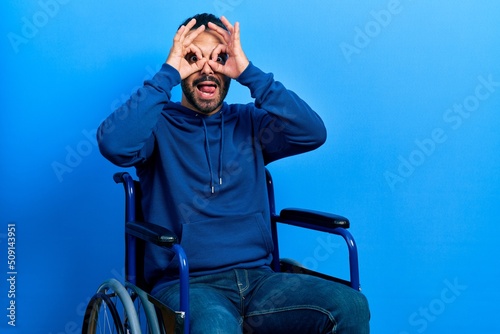 Handsome hispanic man with beard sitting on wheelchair doing ok gesture like binoculars sticking tongue out, eyes looking through fingers. crazy expression.