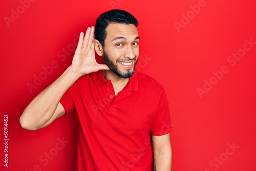 Hispanic man with beard wearing casual red t shirt smiling with hand over ear listening an hearing to rumor or gossip. deafness concept.