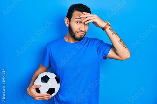 Hispanic man with beard holding soccer ball worried and stressed about a problem with hand on forehead, nervous and anxious for crisis