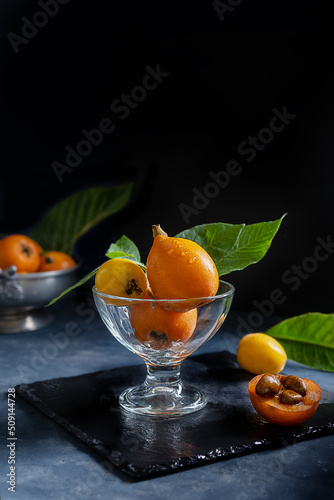Yellow and orange Medlar fruits in glass cup over dark grey background. Chiaroscuro mood photo