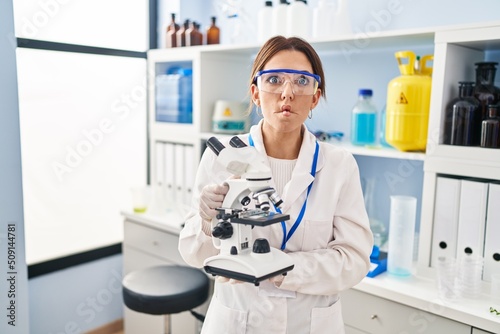 Young brunette woman working at scientist laboratory with microscope making fish face with mouth and squinting eyes  crazy and comical.