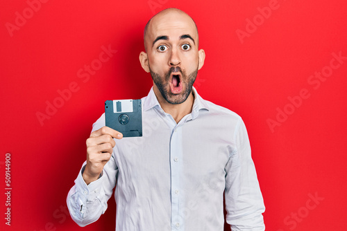 Young bald man holding floppy disk scared and amazed with open mouth for surprise, disbelief face