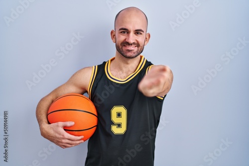 Young bald man with beard wearing basketball uniform holding ball smiling cheerful offering palm hand giving assistance and acceptance.