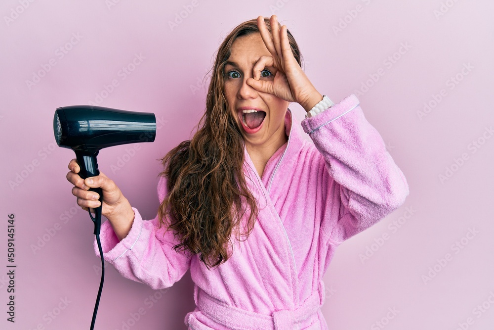 Young blonde woman wearing bathrobe using hair dryer smiling happy doing ok sign with hand on eye looking through fingers Young blonde woman wearing bathrobe using hair dryer smiling happy doing ok sign with hand on eye looking through fingers