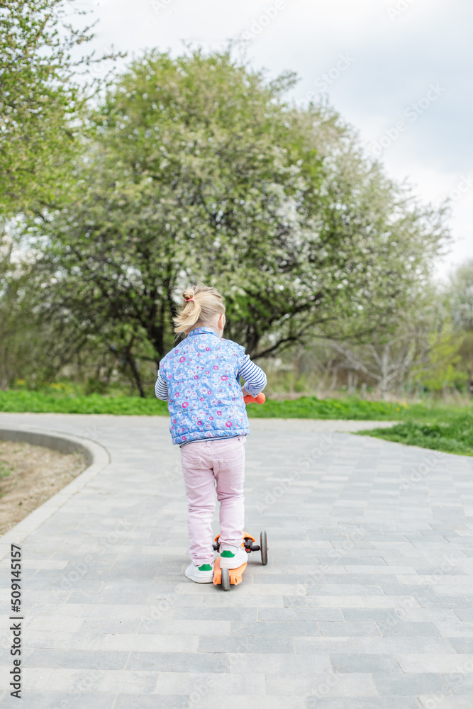 Little toddler girl learns to ride kick scooter in city park on spring day. Little toddler girl learns to ride kick scooter in city park on spring day.