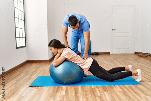 Latin man and woman wearing physiotherapist uniform having rehab session using fit ball at rehab center