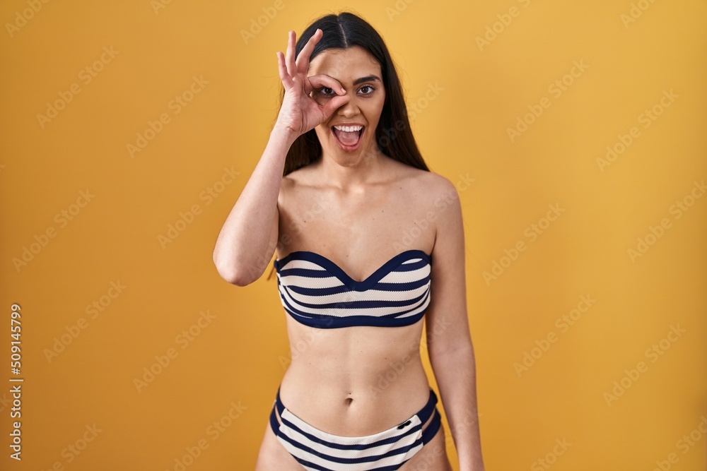 Young brunette woman wearing bikini over yellow background doing ok gesture with hand smiling, eye looking through fingers with happy face. Young brunette woman wearing bikini over yellow background doing ok gesture with hand smiling, eye looking through fingers with happy face.