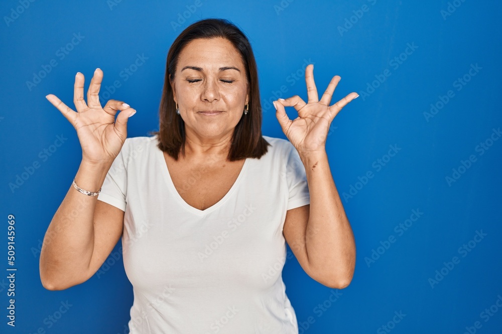 Hispanic mature woman standing over blue background relax and smiling with eyes closed doing meditation gesture with fingers. yoga concept. Hispanic mature woman standing over blue background relax and smiling with eyes closed doing meditation gesture with fingers. yoga concept.