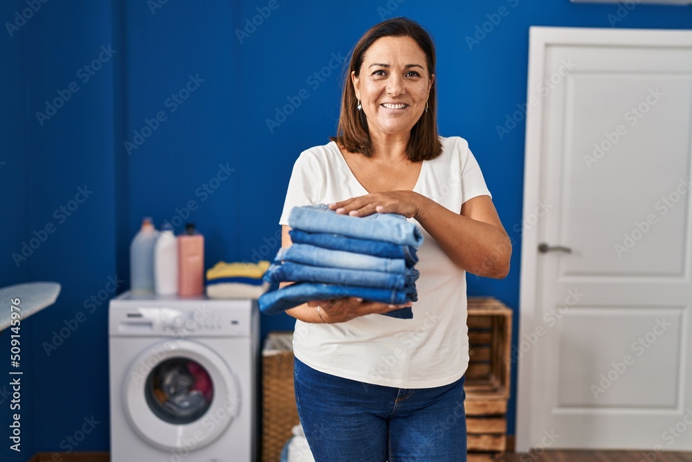 Middle age hispanic woman smiling confident holding folded jeans at laundry room Middle age hispanic woman smiling confident holding folded jeans at laundry room