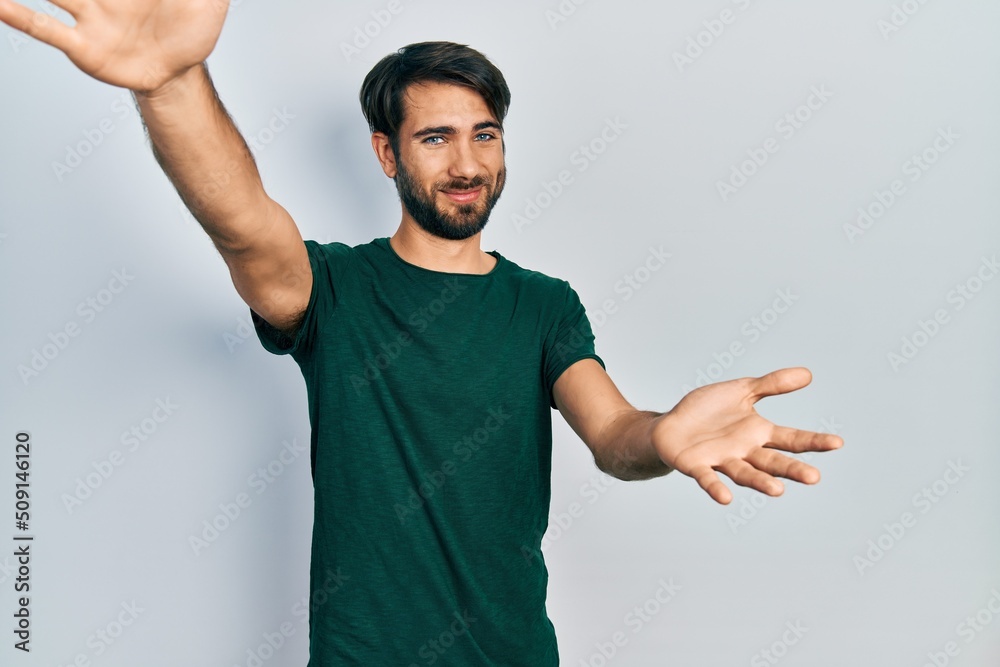 Fototapeta premium Young hispanic man wearing casual white tshirt looking at the camera smiling with open arms for hug. cheerful expression embracing happiness.