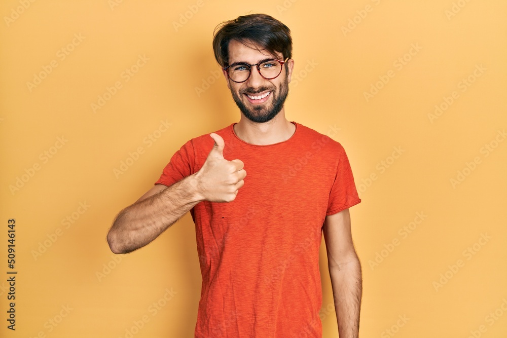 Young hispanic man wearing casual clothes and glasses doing happy thumbs up gesture with hand. approving expression looking at the camera showing success. Young hispanic man wearing casual clothes and glasses doing happy thumbs up gesture with hand. approving expression looking at the camera showing success.