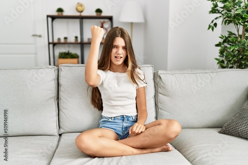 Young brunette teenager sitting on the sofa at home angry and mad raising fist frustrated and furious while shouting with anger. rage and aggressive concept.
