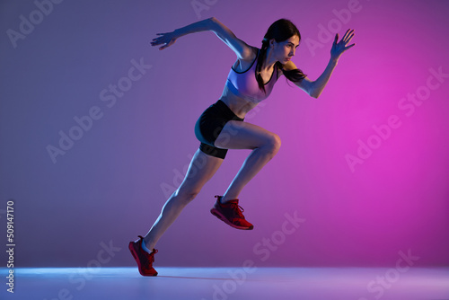 One young muscular girl, female runner or jogger training isolated on pink-blue background in neon light. Sport, track-and-field athletics, competition and active lifestyle concept