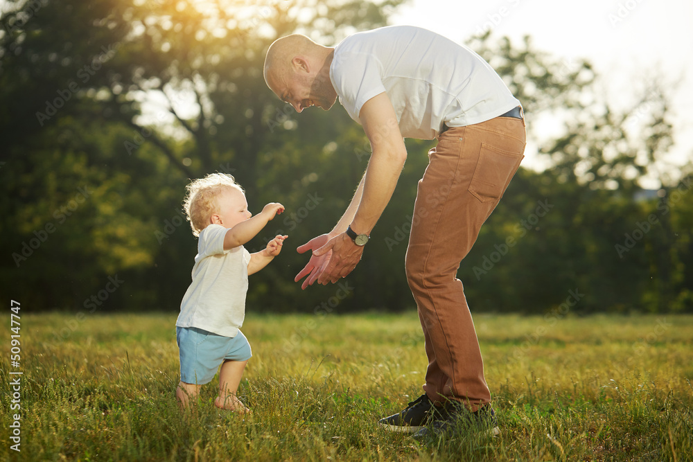 First baby steps. A young father helps a baby boy walk on the grass in the park. Parental care and support