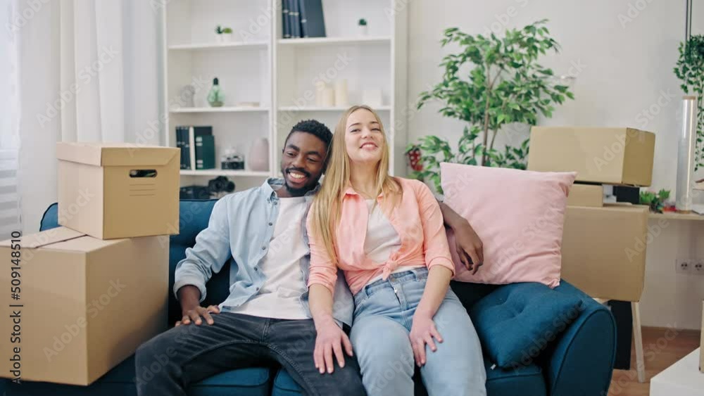 Happy interracial couple sitting on sofa and smiling, buying new home, moving in