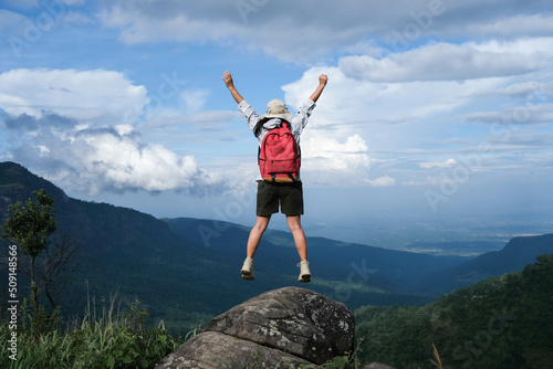 Happy female hiker jumps on a rock with arms outstretched against mountain valley with blue sky. Happy woman enjoying nature in the mountains and looking at sky with raised hands. Freedom concept