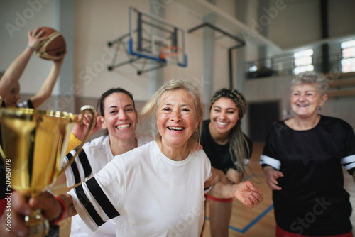 Group of young and old women  basketball team players  in gym with trophy celebrating victory.