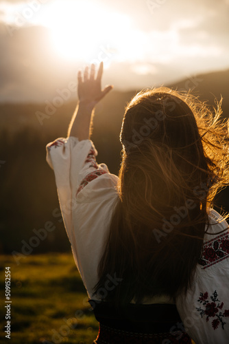 Ukrainian woman rise hand to sun in Carpathians mountains in an embroidered shirt ( vyshyvanka ) from back on sunset. Stand with Ukraine. Symbol of freedom and unconquered photo