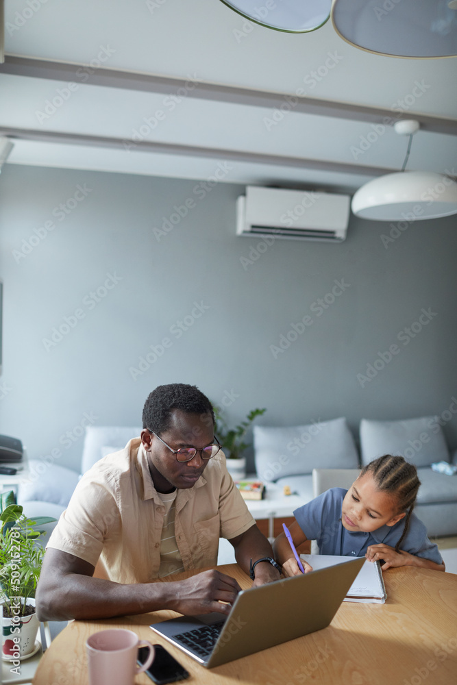 Vertical portrait of African American man working from home and using laptop with cute daughter drawing by his side Vertical portrait of African American man working from home and using laptop with cute daughter drawing by his side