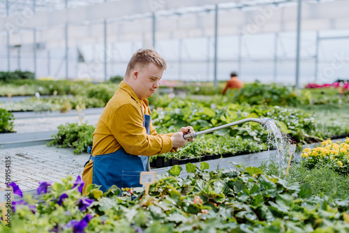 Happy young employee with Down syndrome working in garden centre, watering plants with a shower head and hose. photo