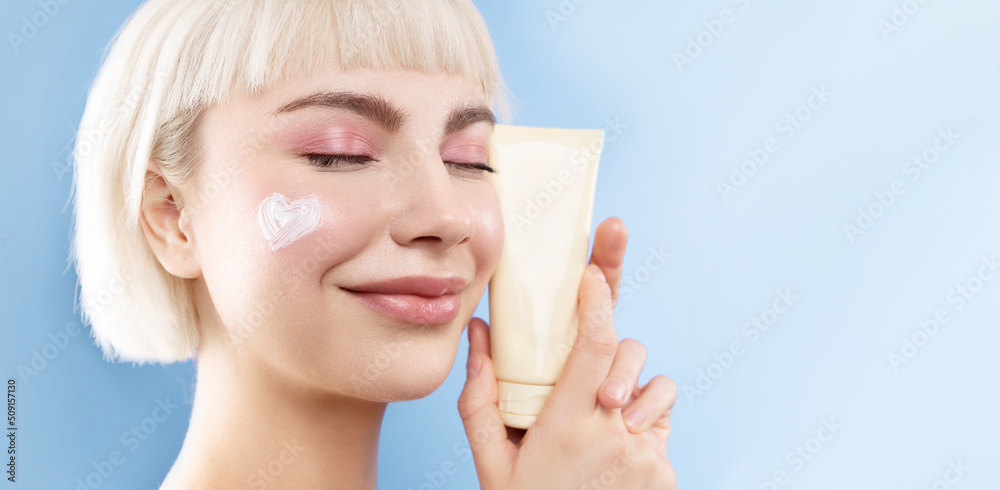 Satisfied smiling blond young girl demonstrating moisturizing face cream mock up plastic jar or tube with copy space. Pretty stylish female model with closed eyes having fun with skincare cosmetics. Satisfied smiling blond young girl demonstrating moisturizing face cream mock up plastic jar or tube with copy space. Pretty stylish female model with closed eyes having fun with skincare cosmetics.
