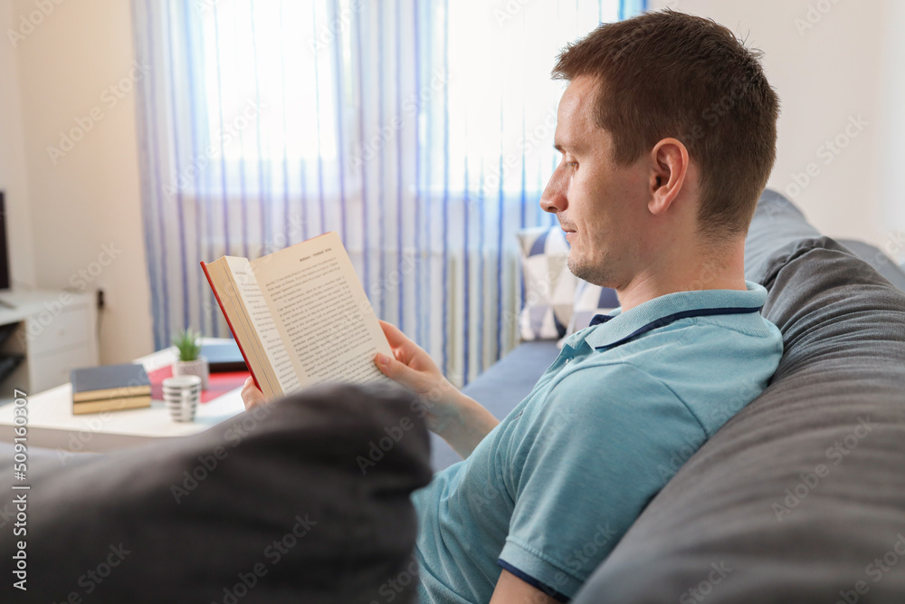 Young man reading book at home, preparing for exams at university, enjoying novels at leisure time. Caucasian man sitting on the sofa bed reading the book study learning. Young man reading book at home, preparing for exams at university, enjoying novels at leisure time. Caucasian man sitting on the sofa bed reading the book study learning.