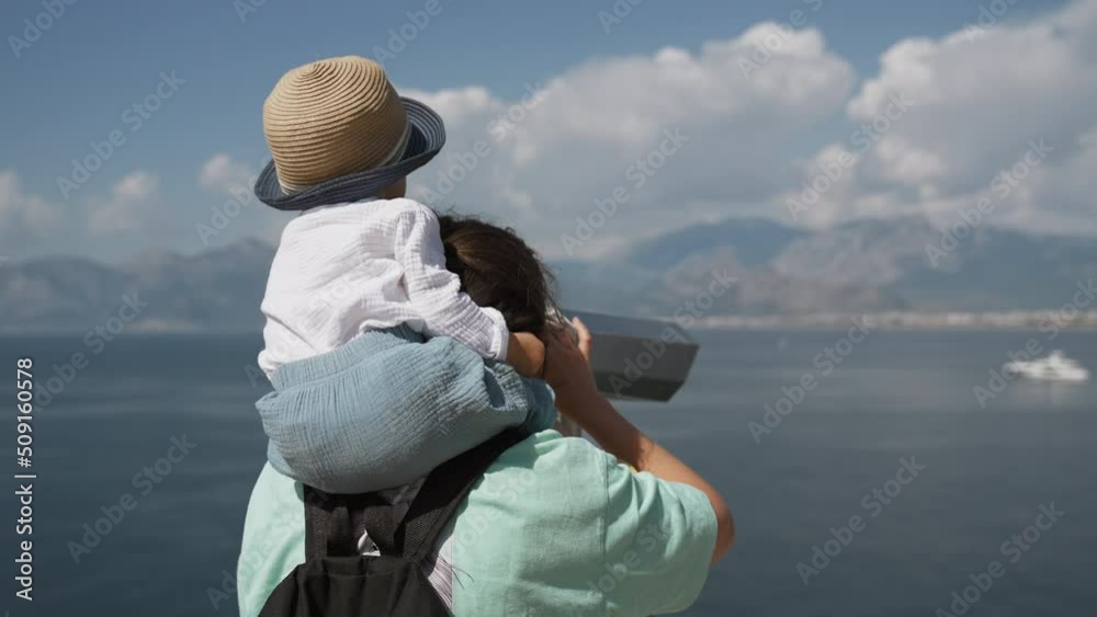 Mom with son on shoulders turns binoculars to look at vessel sailing in sea against mountains. Woman and toddler enjoy vacation in seaside town closeup Mom with son on shoulders turns binoculars to look at vessel sailing in sea against mountains. Woman and toddler enjoy vacation in seaside town closeup