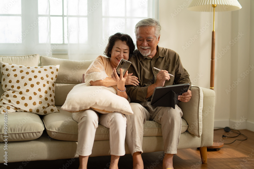 Social distancing or new normal concept.Senior asian couple sitting on sofa in home playing smartphone and video conference with grandchild.They are smiling to spend their time together with happiness Social distancing or new normal concept.Senior asian couple sitting on sofa in home playing smartphone and video conference with grandchild.They are smiling to spend their time together with happiness