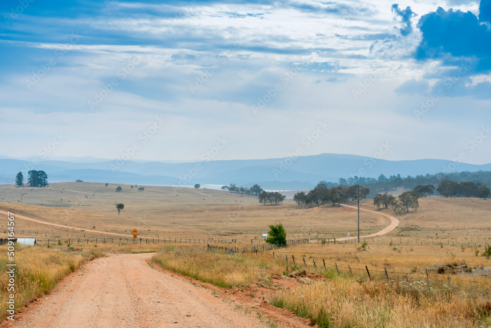Obraz premium Country unsealed gravel dirt road surrounded by farms and fields in Australia. Mountains on the horizon. Road trip rural travel