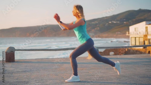 Slow motion shot young female athlete practicing lower body exercise. She doing forward moving lunge against amazing ocean mountain scenery. Caucasian fitness female doing lunges workout training
