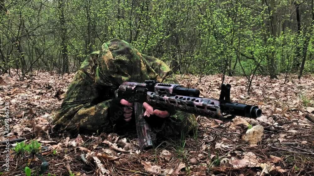 Shooter sniper hides on the ground behind a tree in the woods. Rifleman sniper in camouflage clothing camouflages in a firing position among the dry grass. Shooter sniper hides on the ground behind a tree in the woods. Rifleman sniper in camouflage clothing camouflages in a firing position among the dry grass.