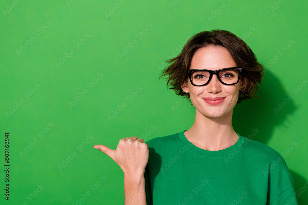 Photo of cute adorable woman wear casual t-shirt eyeglasses pointing thumb empty space isolated green color background Photo of cute adorable woman wear casual t-shirt eyeglasses pointing thumb empty space isolated green color background