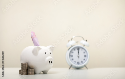 Banknotes are wearing white piggy banks and a pile of coins and an alarm clock on a white table, business, investment, finance, and money savings for future concepts