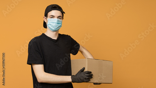 Male delivery man in a black t-shirt, cap, protective mask and gloves with a cardboard box on a yellow background. The concept of delivery under covid-19 restrictions.