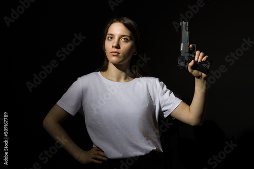 young woman with long hair in a white t-shirt holding a gun in her hand on a black background